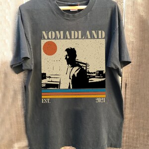 May include: Dark gray t-shirt with the word "NOMADLAND" above a graphic of a person and a sun. Below the graphic are horizontal stripes in orange, teal, and yellow. The text "EST. 2021" is below the stripes.