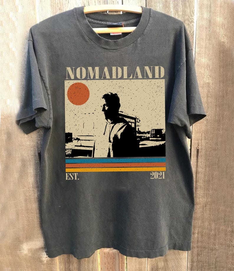 May include: Dark gray t-shirt with the word "NOMADLAND" above a graphic of a person and a sun. Below the graphic are horizontal stripes in orange, teal, and yellow. The text "EST. 2021" is below the stripes.