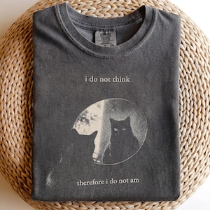 May include: A charcoal gray crew neck t-shirt featuring a graphic print of a cat silhouette and a cat face inside a circular moon-like shape. The text "i do not think" is printed above the graphic, and "therefore i do not am" is printed below.