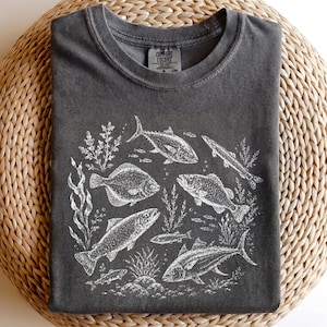 Può includere: Una t-shirt grigio antracite con una stampa grafica bianca di varie specie di pesci e piante acquatiche. La maglietta presenta un girocollo e un'etichetta del marchio Comfort Colors all'interno del colletto.