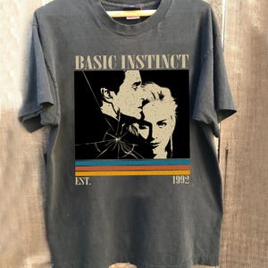 May include: Dark gray t-shirt featuring a graphic from the movie "Basic Instinct." The design includes a black and white image of two people, with the text "BASIC INSTINCT" above and "EST. 1992" below, with orange, blue, and yellow stripes.