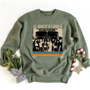 May include: Olive green sweatshirt featuring a graphic from the movie "Almost Famous." The graphic includes the movie title, a tour bus, and a group of people. The design has a retro style with the text "Almost Famous-Tour 73" and the year "2000".