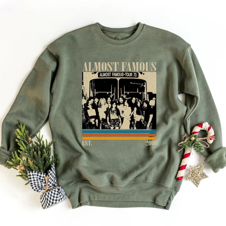 May include: Olive green sweatshirt featuring a graphic from the movie "Almost Famous." The graphic includes the movie title, a tour bus, and a group of people. The design has a retro style with the text "Almost Famous-Tour 73" and the year "2000".