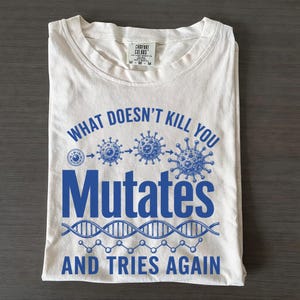 Op de afbeelding: Een crèmekleurig t-shirt met korte mouwen en een blauwe grafische print. Het ontwerp bevat de tekst "WHAT DOESN'T KILL YOU Mutates AND TRIES AGAIN", samen met illustraties van een cel, virusdeeltjes en een DNA-dubbelhelixstreng.