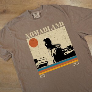 May include: A taupe-colored t-shirt featuring a graphic with the word "NOMADLAND" above a silhouette of a person. The graphic includes a red circle, and horizontal stripes in blue, orange, and yellow. The text "EST. 2021" is also present.