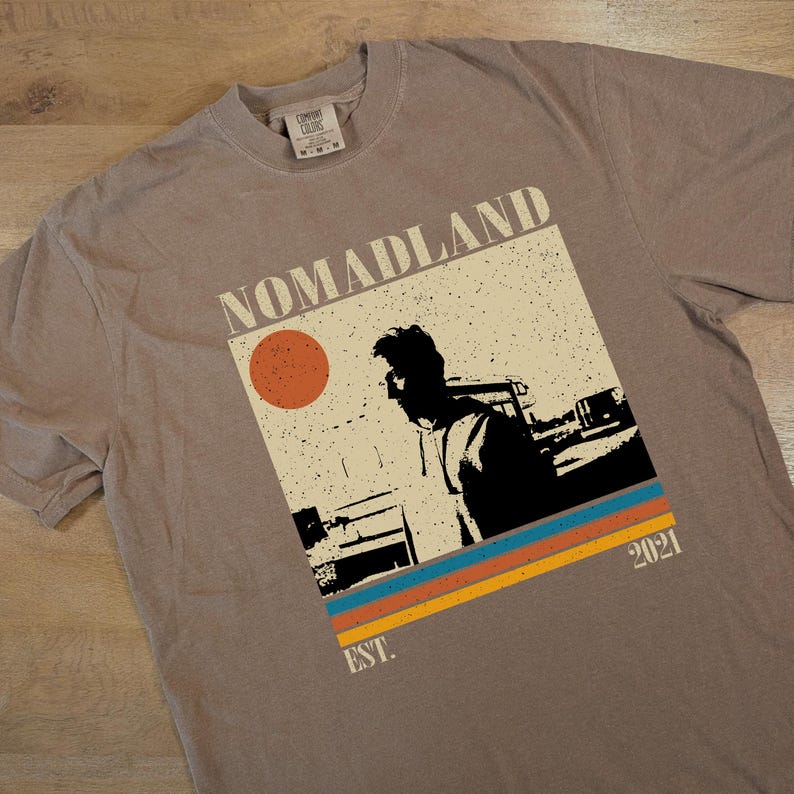 May include: A taupe-colored t-shirt featuring a graphic with the word "NOMADLAND" above a silhouette of a person. The graphic includes a red circle, and horizontal stripes in blue, orange, and yellow. The text "EST. 2021" is also present.