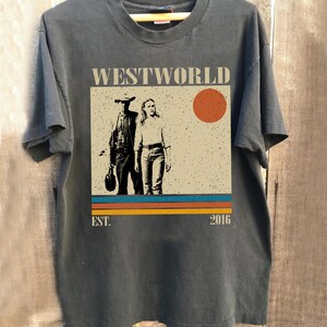 Op de afbeelding: Donkergrijs t-shirt met een afbeelding van de tv-serie "Westworld". Het ontwerp toont twee figuren, een rode zon en de tekst "WESTWORLD" erboven. Daaronder horizontale strepen en de tekst "EST. 2016."