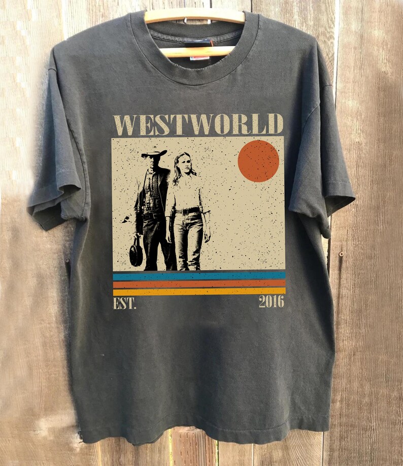 Op de afbeelding: Donkergrijs t-shirt met een afbeelding van de tv-serie "Westworld". Het ontwerp toont twee figuren, een rode zon en de tekst "WESTWORLD" erboven. Daaronder horizontale strepen en de tekst "EST. 2016."