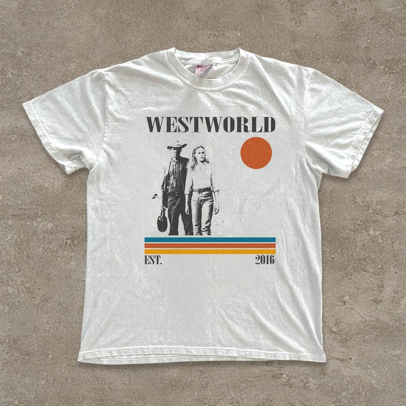 Op de afbeelding: Wit t-shirt met een grafische print. Het ontwerp bevat het woord "WESTWORLD", een zwart-wit afbeelding van twee figuren, een oranje cirkel en de tekst "EST. 2016". Onder de afbeelding bevinden zich horizontale strepen in blauw, oranje en geel.