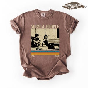 Puede incluir: Camiseta color espresso con un gráfico que muestra a dos personas sentadas en escalones, con las palabras "NORMAL PEOPLE" arriba y "EST. 2020" abajo. El gráfico tiene un diseño retro con rayas azules, naranjas y amarillas.