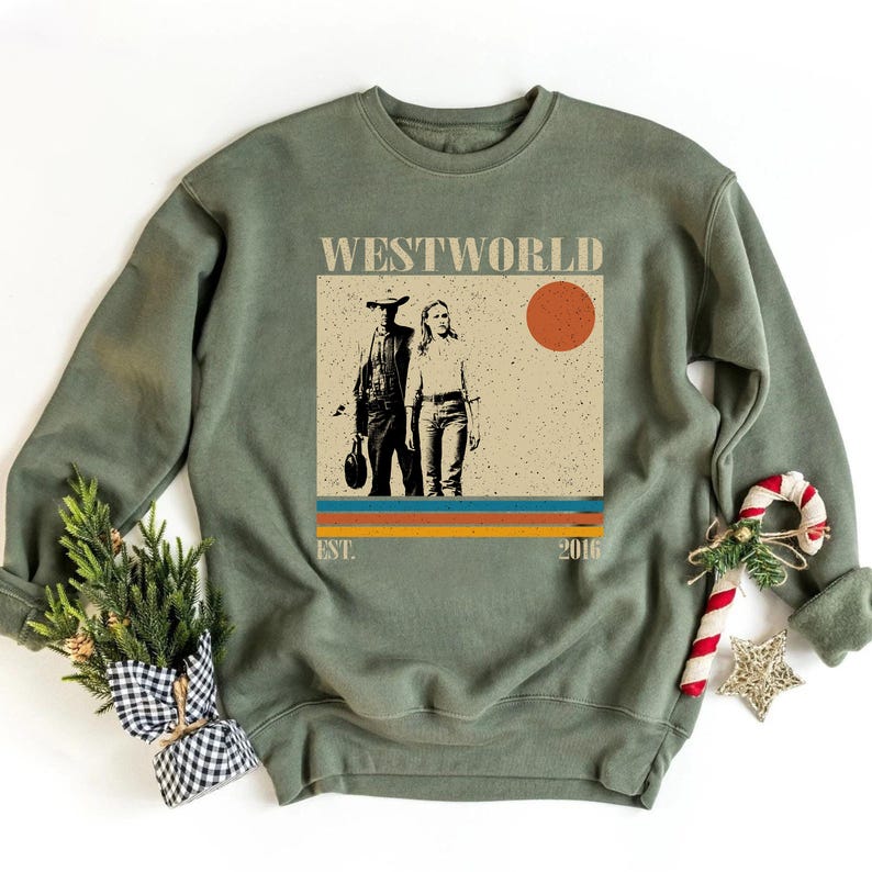 Op de afbeelding: Olijfgroene sweatshirt met een afbeelding van de tv-serie "Westworld". Het ontwerp bevat twee figuren, de titel van de show, een zon en het jaar 2016. De sweatshirt heeft een ronde hals en lange mouwen.