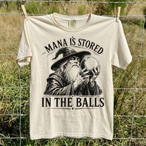 Puede incluir: Una camiseta de manga corta de color crema con un estampado gráfico negro de un mago barbudo con sombrero puntiagudo que sostiene una bola de cristal. El texto sobre el mago dice MANA IS STORED y el texto debajo dice IN THE BALLS.
