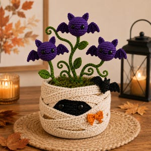Peut inclure: Un pot de plante au crochet enveloppé de fil beige pour ressembler à une momie, avec un petit nœud orange et une chauve-souris noire. Trois tiges vertes émergent du haut, chacune surmontée d'une chauve-souris violette avec des yeux noirs.
