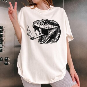 Könnte beinhalten: Ein cremefarbenes T-Shirt mit Rundhalsausschnitt und einer detaillierten schwarz-weißen Grafikillustration einer Schlange mit geöffnetem Maul, die scharfe Giftzähne zeigt und eine brennende, rauchende Zigarette im Kiefer hält.