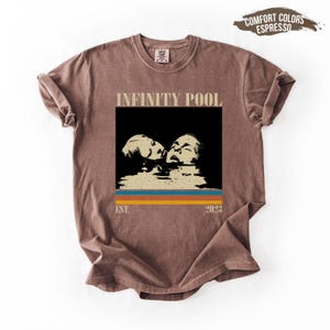 May include: Espresso-colored t-shirt featuring a graphic with the words "INFINITY POOL" above a black square with two faces in water. Below the square are horizontal stripes in blue, orange, and yellow. The text "EST. 2023" is below the stripes.