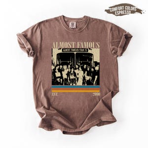 May include: Espresso-colored t-shirt featuring a graphic from the movie "Almost Famous." The design includes the text "Almost Famous," "Almost Famous Tour 73," and the years "Est. 2000." The graphic shows a group of people in front of two buses, with a retro color scheme.