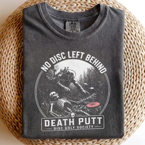 Pode incluir: Uma t-shirt cinzenta escura de gola redonda com uma impressão gráfica branca de um mergulhador e um esqueleto debaixo de água. O texto diz NO DISC LEFT BEHIND e DEATH PUTT DISC GOLF SOCIETY. Um pequeno disco cor-de-rosa é visível perto da mão.