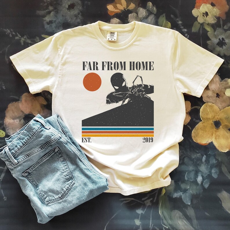 May include: Cream t-shirt featuring the text "FAR FROM HOME" and a superhero graphic. Below the graphic are the words "EST. 2019" and horizontal stripes. Light blue denim jeans are folded beside the shirt.
