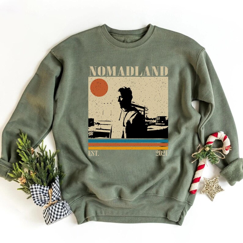 May include: Sage green sweatshirt with a graphic featuring the word "NOMADLAND" above a retro design of a person and a sun. Below the design are horizontal stripes in orange, yellow, and blue. The text "EST. 2021" is below the stripes.