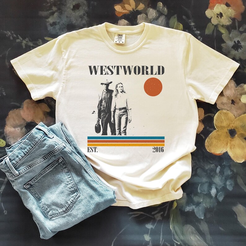 Op de afbeelding: Cr&egrave;mekleurig T-shirt met de tekst "WESTWORLD" boven een afbeelding van twee figuren en een rode cirkel. Onder de afbeelding staan de woorden "EST. 2016" boven een gestreept ontwerp. Een lichtblauwe jeans ligt naast het shirt.