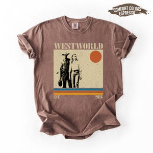 Op de afbeelding: Bruin T-shirt met een afbeelding van de tv-serie "Westworld". Het ontwerp toont twee figuren, een zon en de tekst "Westworld" erboven. Het shirt heeft ook de tekst "EST. 2016" eronder.