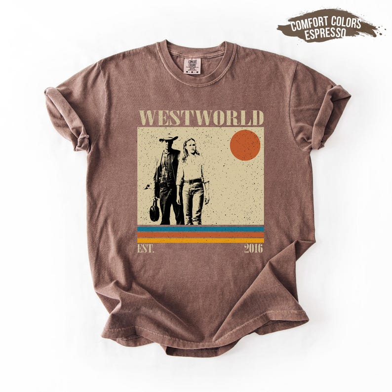 Op de afbeelding: Bruin T-shirt met een afbeelding van de tv-serie "Westworld". Het ontwerp toont twee figuren, een zon en de tekst "Westworld" erboven. Het shirt heeft ook de tekst "EST. 2016" eronder.