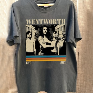 May include: Dark gray t-shirt featuring a graphic of a group of people with the word "WENTWORTH" above them. Below the image are horizontal stripes in orange, blue, and yellow, with "EST. 2013" below the stripes. The shirt is hanging on a wooden hanger.