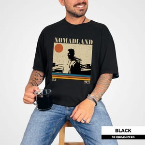 May include: Black t-shirt with a retro-style graphic that reads "NOMADLAND" above a silhouette of a person and a sunset. The shirt also has the text "EST. 2021" below the graphic. The shirt is worn by a person.