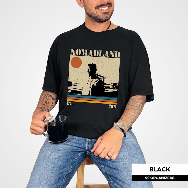 May include: Black t-shirt with a retro-style graphic that reads "NOMADLAND" above a silhouette of a person and a sunset. The shirt also has the text "EST. 2021" below the graphic. The shirt is worn by a person.