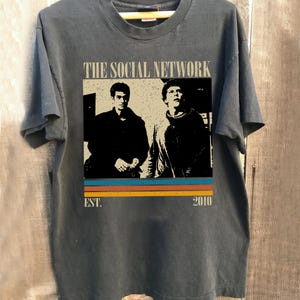 May include: Dark gray t-shirt featuring a graphic from "The Social Network" film. The design includes a sepia-toned image of two men, the film title, and the text "EST. 2010" with orange, yellow, and blue stripes. The shirt is hanging on a wooden hanger.