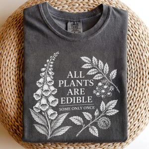 May include: A charcoal gray crew neck t-shirt featuring a white botanical illustration of foxglove and berry branches. The text printed in white reads: ALL PLANTS ARE EDIBLE SOME ONLY ONCE. The shirt has a Comfort Colors brand label at the neck.