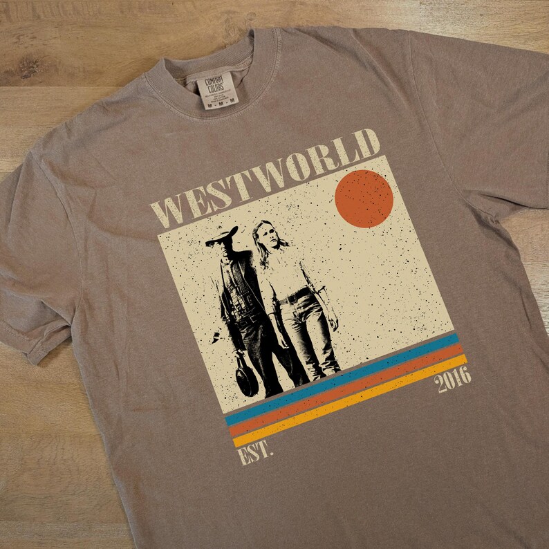 Op de afbeelding: Een taupekleurig t-shirt met een afbeelding van de serie "Westworld". Het ontwerp bevat twee figuren, een zon en de tekst "WESTWORLD" en "2016". Het shirt heeft een vintage, distressed look.