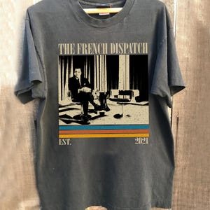 Puede incluir: Camiseta gris oscuro con un diseño gráfico de la película "The French Dispatch". El diseño incluye una figura sentada, el título de la película y el año 2021, con rayas retro. La camiseta se muestra en una percha de madera.