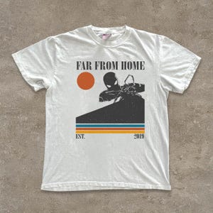 May include: White t-shirt with a graphic design. The design includes the text "FAR FROM HOME", an orange sun, and a character silhouette. Below the graphic are the words "EST. 2019" and three horizontal stripes.