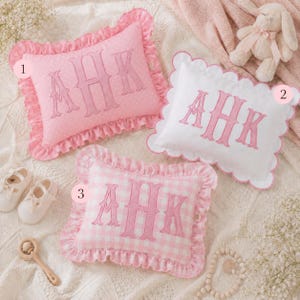 May include: Three rectangular decorative pillows featuring pink embroidered AHK monograms. Pillow 1 is pink with white polka dots and a ruffled edge. Pillow 2 is white with a scalloped edge. Pillow 3 is pink and white gingham with a ruffled edge.