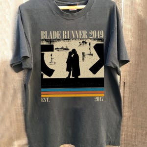 May include: Dark gray t-shirt featuring the text "BLADE RUNNER 2049" above a silhouette of two figures. Below the figures are horizontal stripes in orange, blue, and yellow. The text "EST. 2017" is printed below the stripes.