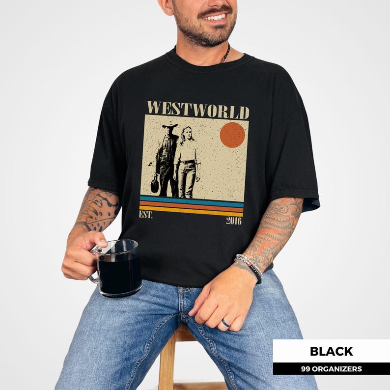 Op de afbeelding: Zwart T-shirt met een afbeelding van de tv-serie "Westworld". Het ontwerp toont twee figuren, een zon en de tekst "WESTWORLD EST. 2016". Het shirt heeft een ronde hals.