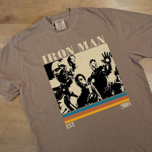 May include: A grey t-shirt with an Iron Man graphic and other characters. The design includes the text "IRON MAN" and "2008" with orange, blue, and yellow stripes. The shirt is made of a soft material. The word "EST." is also visible.