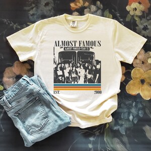 May include: Cream-colored t-shirt with the text "Almost Famous" and a graphic of a tour bus and a group of people. The shirt also has the text "Almost Famous-Tour 73" and "Est. 2000". Light blue denim jeans are folded next to the shirt.