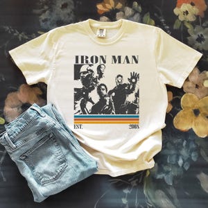 May include: Cream-coloured t-shirt with an Iron Man graphic, the text "IRON MAN" above the image and "EST. 2008" below. The shirt is shown with light blue denim jeans.