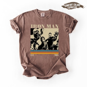 May include: Brown t-shirt with an Iron Man graphic. The design includes the text "IRON MAN" above a rectangular image with multiple figures and the text "EST. 2008" below. The shirt has a crew neck and short sleeves.