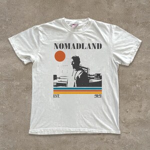 May include: White t-shirt with a graphic design featuring the word "NOMADLAND", a silhouette of a person, an orange sun, and horizontal stripes in blue, orange, and yellow. The text "EST. 2021" is also included.