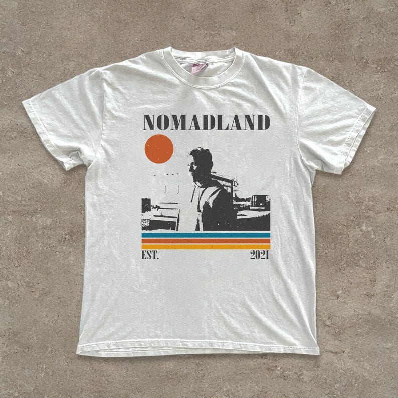 May include: White t-shirt with a graphic design featuring the word "NOMADLAND", a silhouette of a person, an orange sun, and horizontal stripes in blue, orange, and yellow. The text "EST. 2021" is also included.