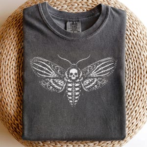 May include: A charcoal gray crew neck t-shirt featuring a white graphic print of a moth with a skull head and skeletal wing patterns. The shirt has a Comfort Colors brand label printed on the inside of the collar area, indicating a size CM.