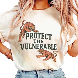 Peut inclure: Un t-shirt à manches courtes de couleur crème avec une impression graphique de deux tigres orange et noirs. Le texte "PROTECT THE VULNERABLE" est imprimé en lettres capitales vert foncé entre les deux tigres, l'un au-dessus et l'autre en dessous.