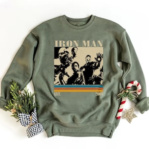 May include: An olive green sweatshirt with an Iron Man graphic. The graphic includes the text "IRON MAN" at the top, and "EST. 2008" at the bottom. The design has a retro style with orange, yellow, and blue stripes.