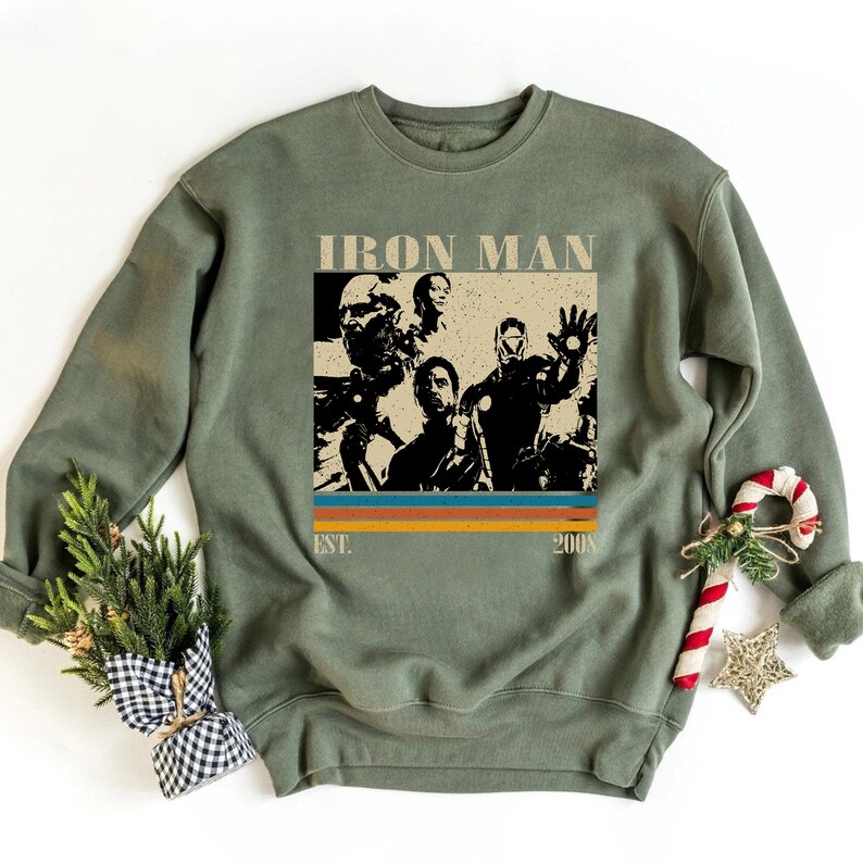 May include: An olive green sweatshirt with an Iron Man graphic. The graphic includes the text "IRON MAN" at the top, and "EST. 2008" at the bottom. The design has a retro style with orange, yellow, and blue stripes.