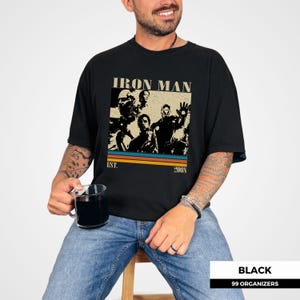 May include: Black t-shirt with a vintage-style graphic featuring the text "IRON MAN" above a collage of characters. Below the graphic are the words "EST. 2008" and a series of horizontal stripes.
