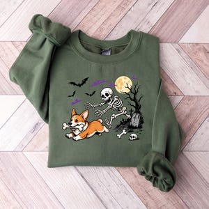 May include: An olive green crewneck sweatshirt featuring a graphic print of a skeleton chasing a Corgi dog that is carrying a bone in its mouth. The scene includes a full moon, bare trees, a tombstone, bats, and purple clouds in the background.