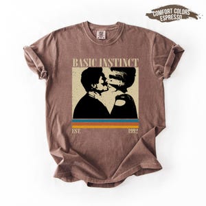 May include: Espresso-colored t-shirt featuring a graphic of two people kissing, with the words "BASIC INSTINCT" above the image. Below the image are the words "EST. 1992" and three horizontal stripes in blue, orange, and yellow.